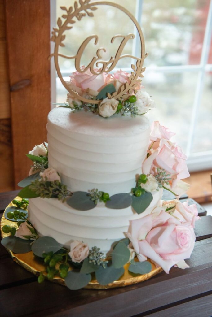 Two-tier white frosted cake adorned with pink roses, greenery, and a gold monogram topper on a wooden table.