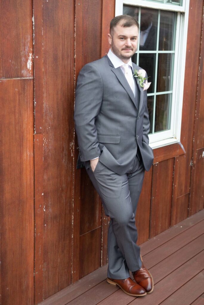 Man in gray suit and brown shoes leans against a wooden wall next to a window, with hands in pockets.