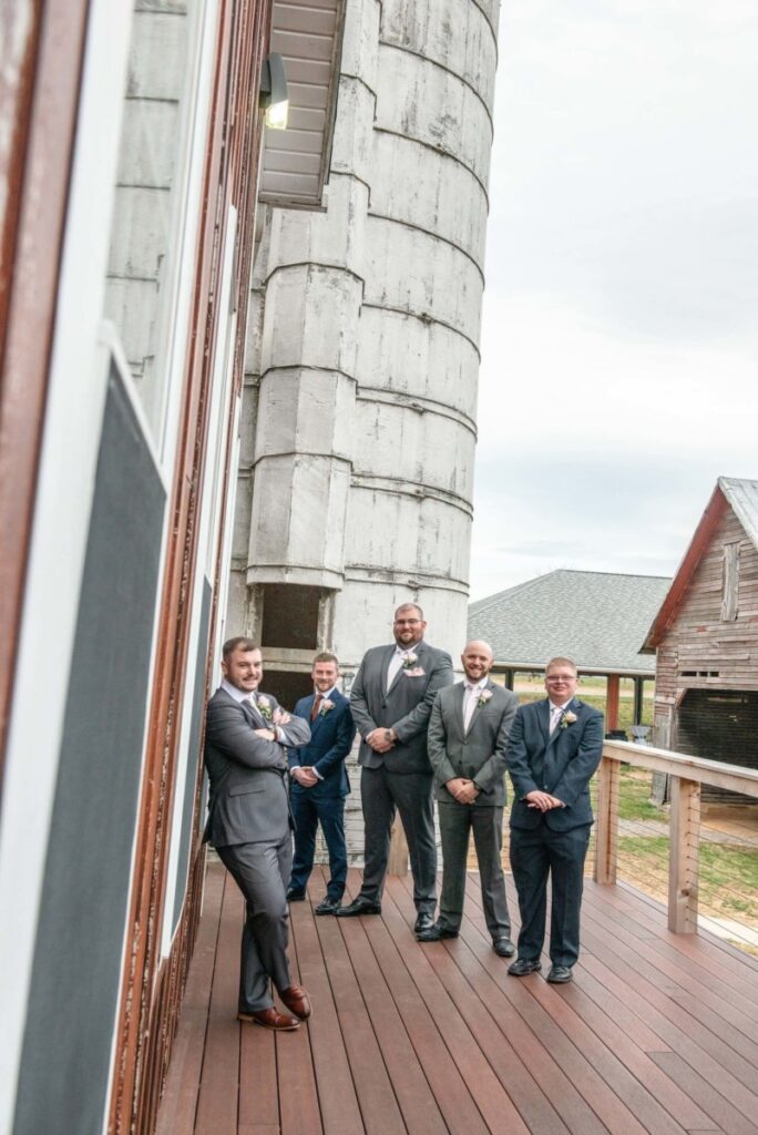 Five men in suits pose on a wooden deck beside a tall, cylindrical structure and rustic buildings.