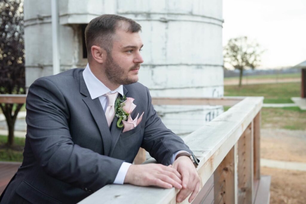 A man in a suit with a boutonniere stands by a wooden railing, looking into the distance. Background includes a silo and some trees.