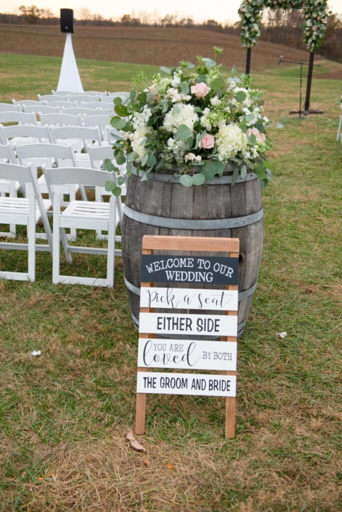 Outdoor wedding setup with white chairs and floral arrangements. A wooden sign reads, "Welcome to our wedding. Pick a seat either side. You are loved by both the groom and bride.