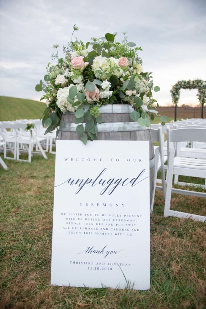 A wooden barrel with a floral arrangement and a sign reading "Welcome to our unplugged ceremony" is placed among rows of white chairs outdoors on a grassy area.