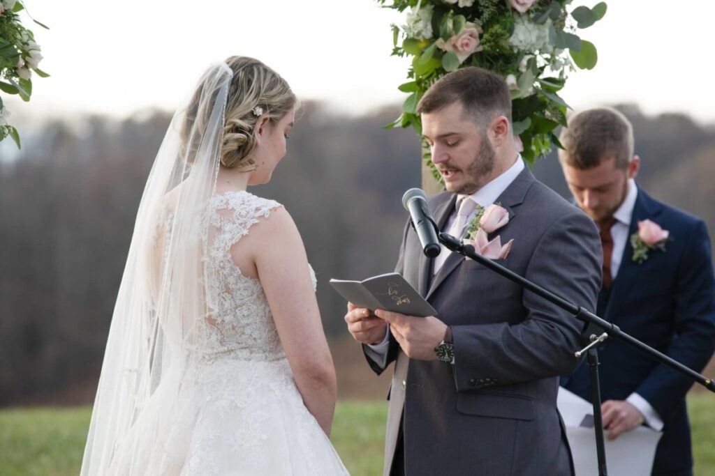 A groom reads vows from a notebook at an outdoor wedding ceremony. The bride, in a lace gown, listens. A groomsman stands in the background, holding papers. Floral decorations are visible.