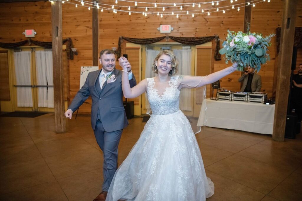 A bride and groom walk hand in hand in a wooden hall. The bride is smiling and holding a bouquet, and the groom is wearing a suit. String lights are hanging above.