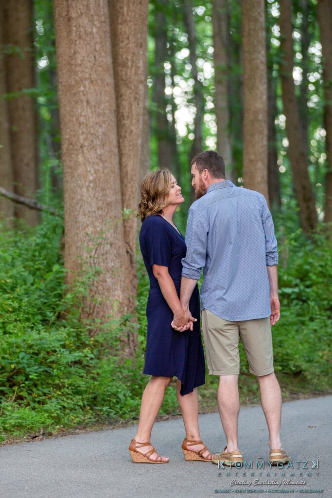 A couple holding hands, with their foreheads touching, stands on a forest path surrounded by tall trees and greenery.