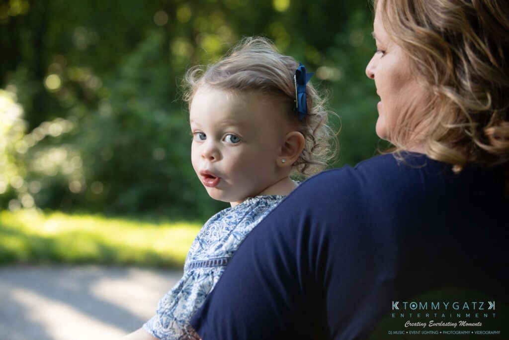 A toddler with a blue bow in her hair being held by an adult outdoors in a park setting.