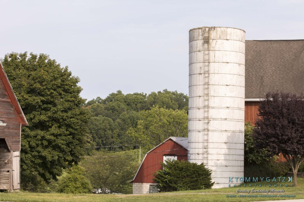 Farm scene with a large white silo, red barn, and trees. The background has a line of lush green trees under a clear sky.