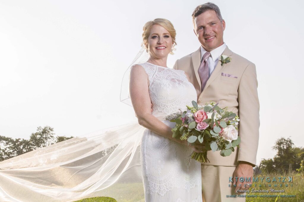 A couple in wedding attire stands outdoors smiling, with the bride holding a bouquet of pink and white flowers.