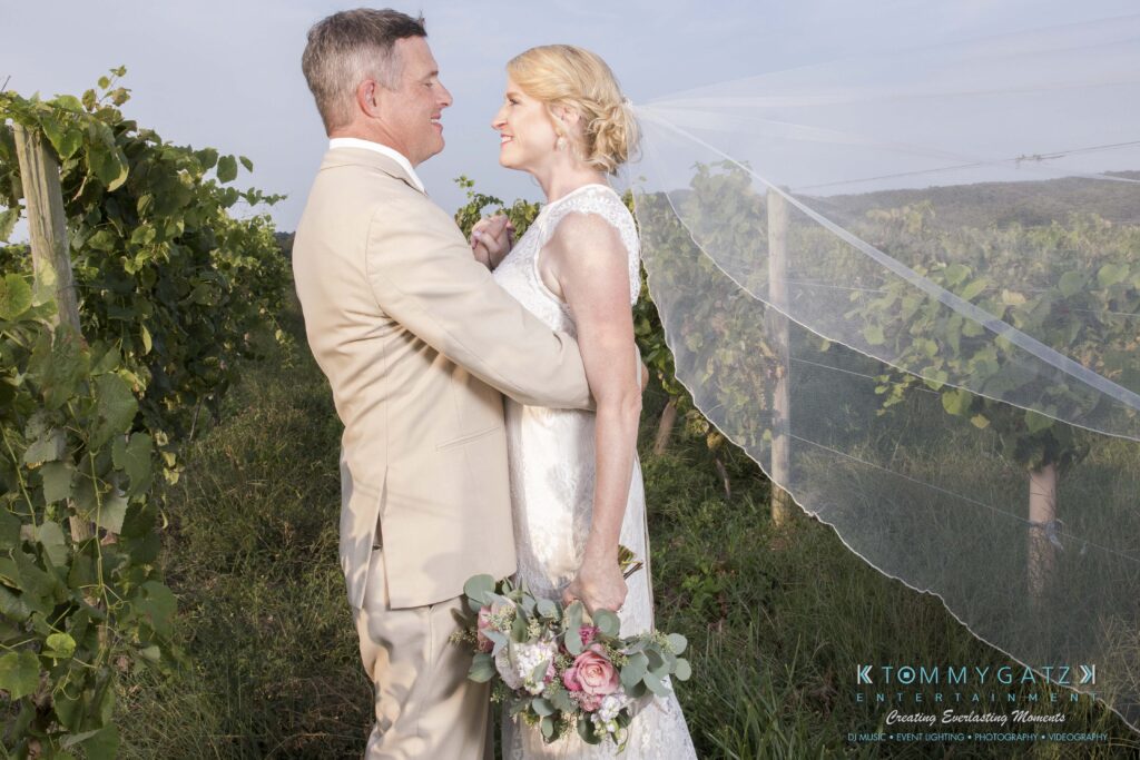 A couple in wedding attire stands smiling in a vineyard, with the bride holding a bouquet and wearing a flowing veil.