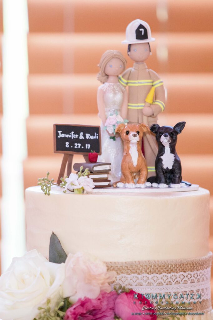 Wedding cake with a firefighter and bride topper, a small chalkboard with "Jennifer & Rudi 8.27.17," two dog figurines, and flowers at the base.