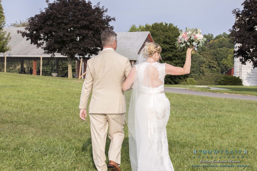 A couple in formal attire walks on grass, with the woman holding a bouquet. They are headed toward a barn and trees in the background.
