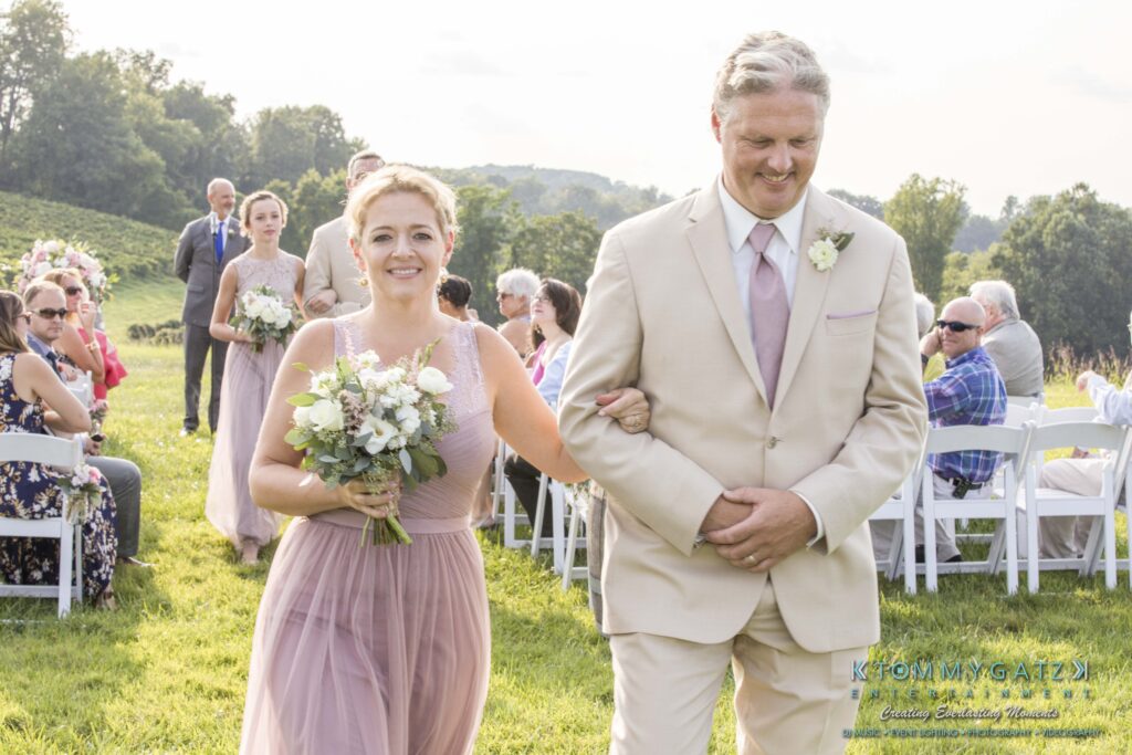 A woman and man in formal attire walk down an outdoor aisle. She holds a bouquet; guests are seated on either side.