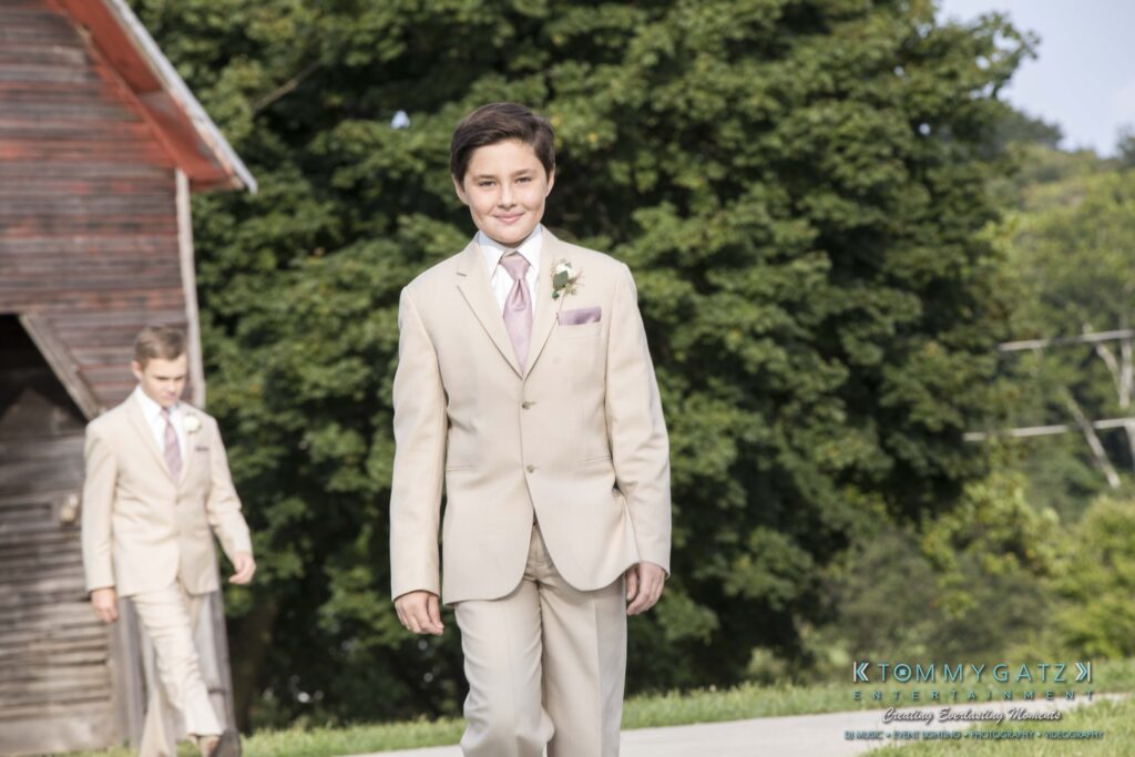 A young boy in a beige suit with a pink tie and boutonnière walks outdoors. Another boy in a similar outfit follows. Trees and a barn are in the background.