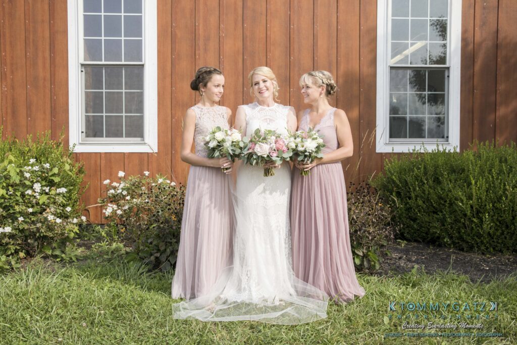 Three women in pastel dresses stand together holding bouquets in front of a wooden building with two windows. The central woman wears a white dress. They are on a grassy area with shrubs.