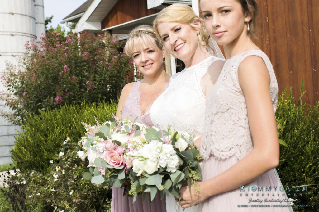 Three women in pastel dresses pose outdoors. The woman in the center holds a bouquet of flowers. A house and silo are visible in the background.