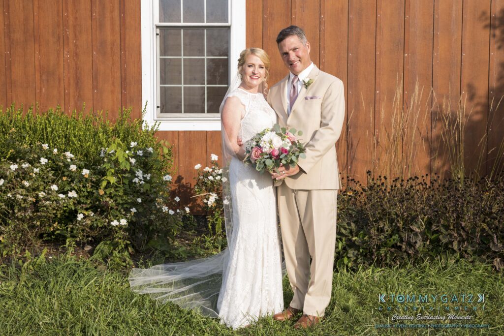 A bride and groom stand smiling in front of a wooden building with a window. The bride holds a bouquet of flowers.