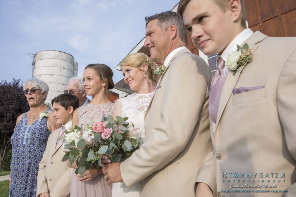 A group of people in formal attire pose for a photo outdoors. A woman holds a bouquet. A silo and wooden structure are in the background.