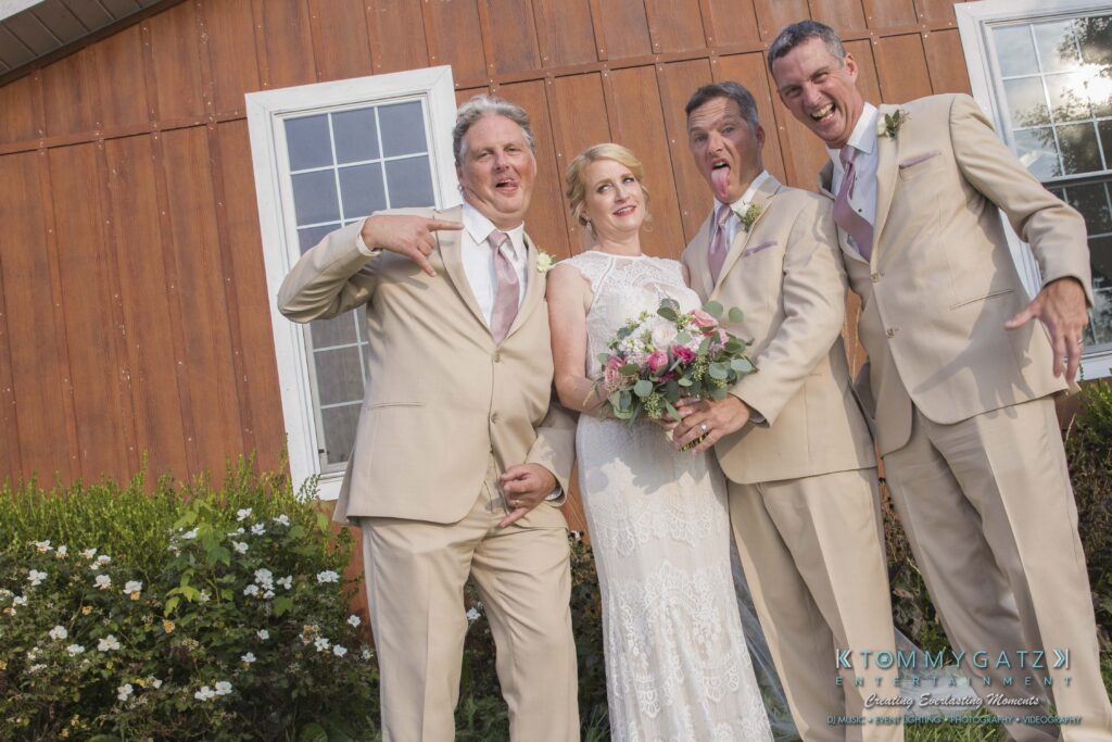 A bride in a white dress stands with three men in beige suits making playful gestures in front of a wooden building with windows and nearby greenery.