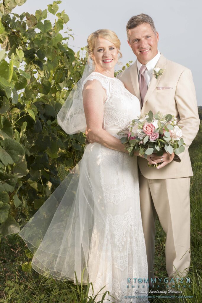 A bride and groom in formal attire stand together in a garden setting, holding a bouquet of flowers.