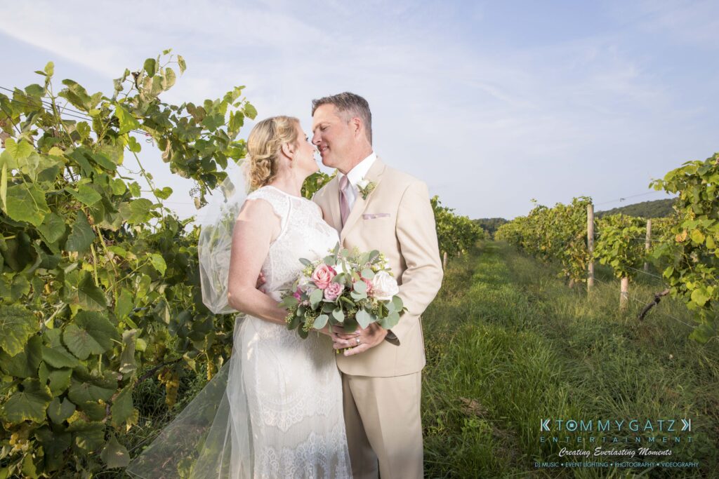A bride and groom embrace and smile at each other in a vineyard, with the bride holding a bouquet of pink and white flowers.