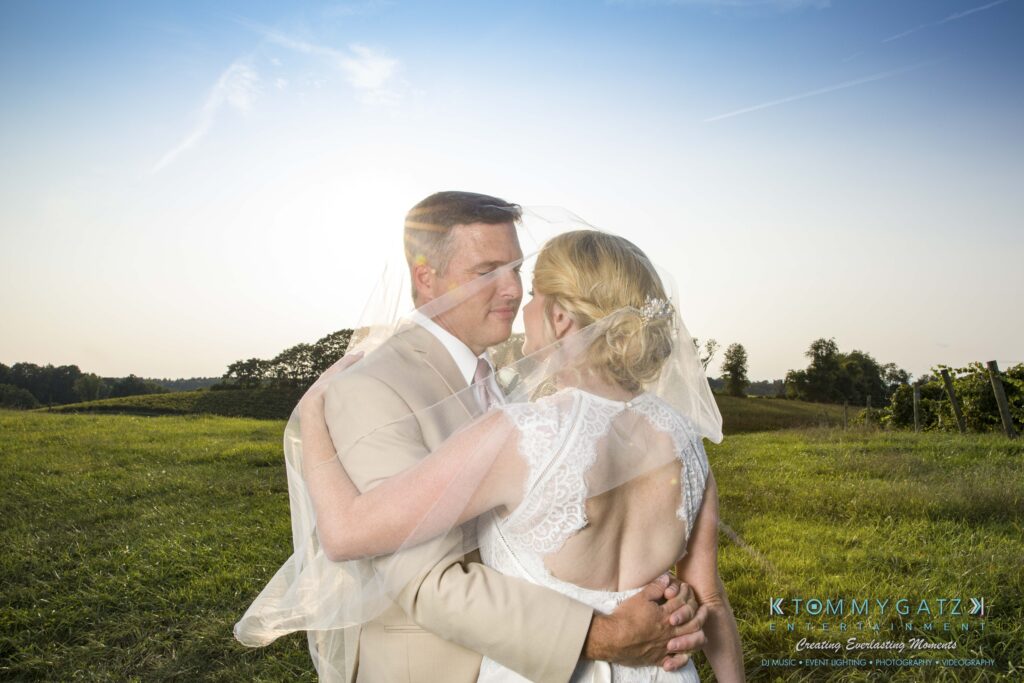 A bride and groom embrace outdoors in a grassy field under a clear sky. The bride's veil is draped over both of them.