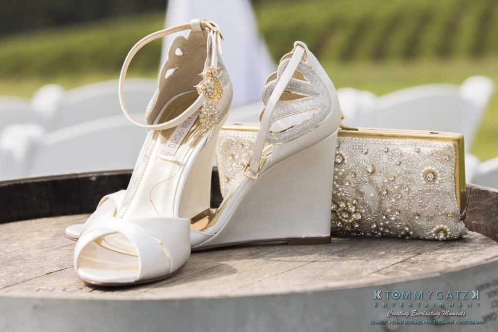 White wedge sandals with decorative ankle straps are placed on a wooden surface next to a gold-embellished clutch.