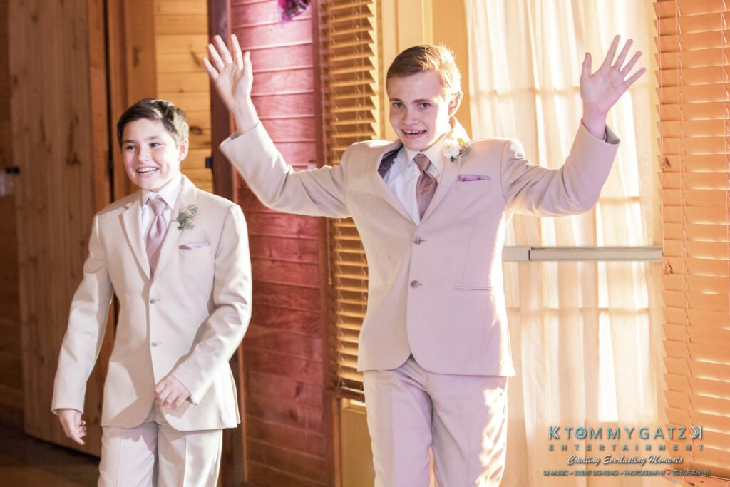 Two boys in beige suits enter a room, the boy on the right raising his arms, both smiling.
