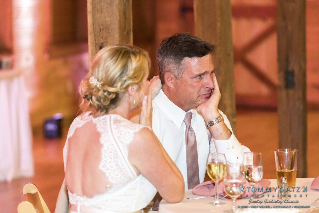 A couple sits at a table during a formal event. The woman has her back turned slightly, while the man looks contemplative. Glasses and dishes are on the table; a wooden structure is behind them.