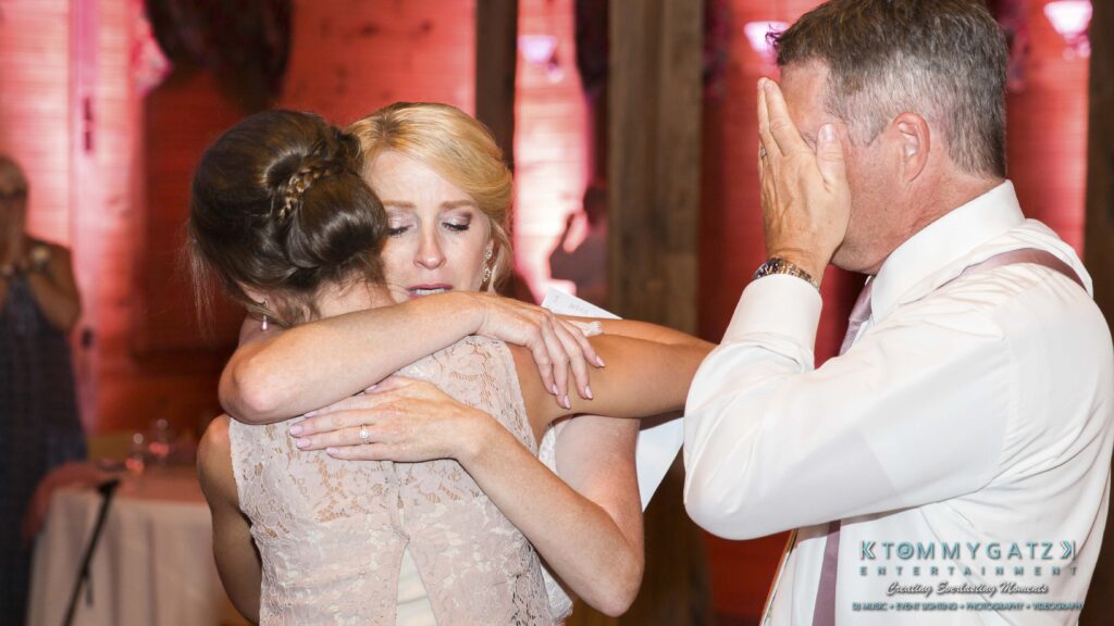 Two women embrace in a hug at a gathering while a man nearby wipes his eyes. A sign in the foreground reads "Tommy Gatz Entertainment.