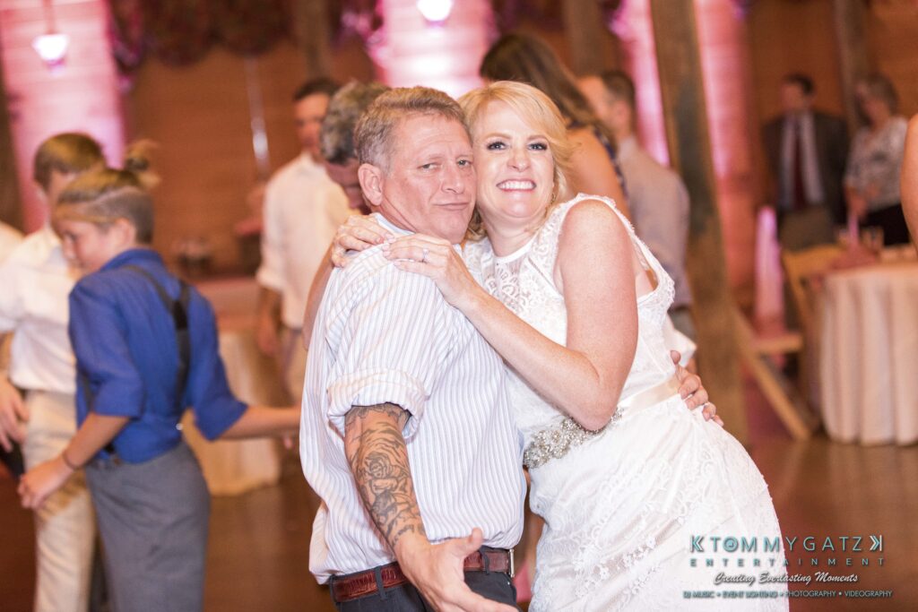 A smiling couple poses closely together in a wedding reception setting. The man has tattoos and wears a striped shirt, while the woman is in a white dress. Guests are visible in the background.