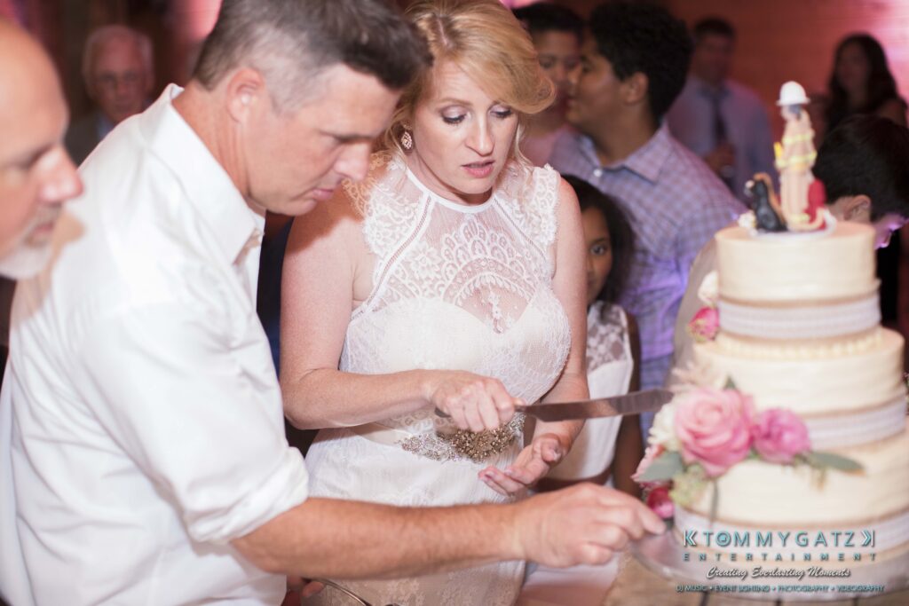 A woman in a white dress cuts a multi-tiered cake with the assistance of a man in a white shirt at a reception.