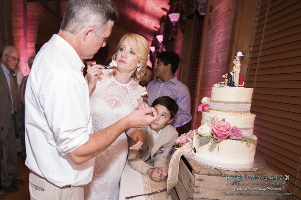 A man and woman stand near a wedding cake with floral decorations. A young boy leans over the table in the background, holding his chin in his hand. Guests are visible in the background.