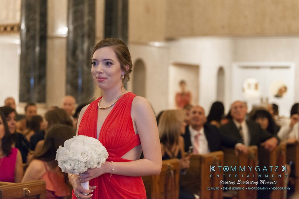 A woman in a red dress holds a bouquet while walking down an aisle in a church, with seated guests watching.