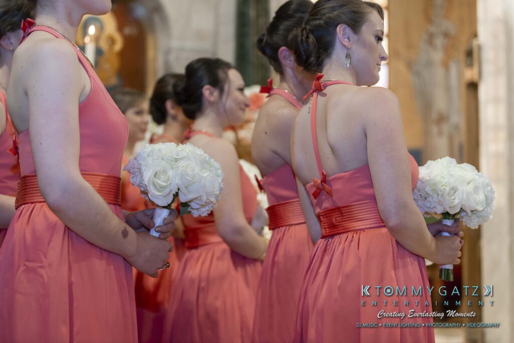 Bridesmaids in coral dresses hold white bouquets, standing in a line at an indoor venue.