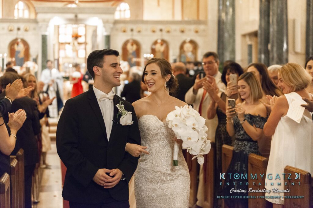A bride and groom walk down the aisle of a church, smiling while guests applaud and take photos.