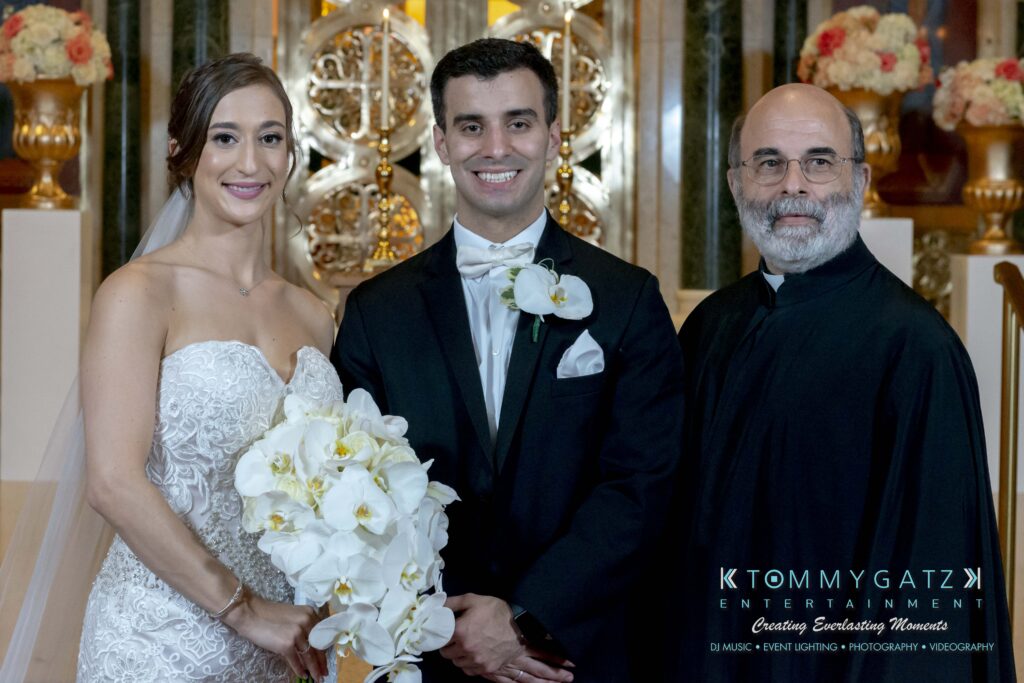 Bride and groom smiling with a man in dark attire, holding a bouquet of white flowers. Elegant interior background.