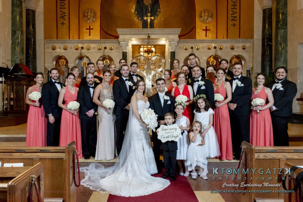 A large wedding party poses inside a church with a bride and groom in the center, surrounded by bridesmaids in coral dresses and groomsmen in black suits.