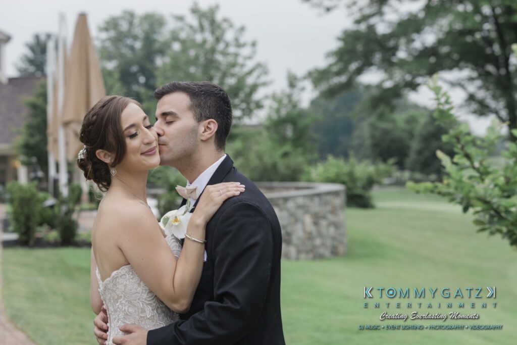 A bride and groom embrace outdoors, with the groom gently kissing the bride's cheek. A stone wall and trees are visible in the background.