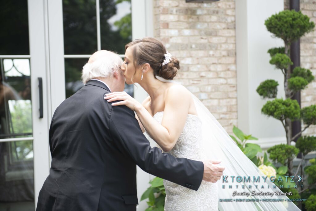 Bride in a white dress kisses an elderly man on the head outdoors near a building, with the groom's name visible on the image.