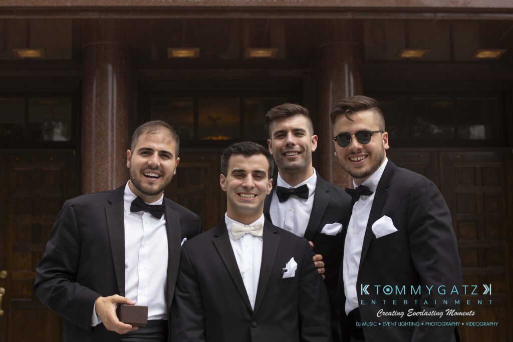 Four men in tuxedos stand together smiling in front of a building entrance.