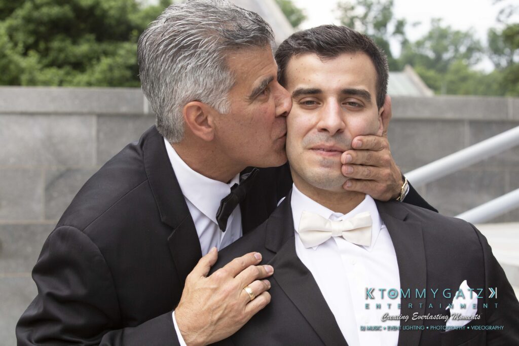 An older man in a tuxedo playfully kissing a younger man's cheek, who seems amused, both wearing formal attire outdoors.