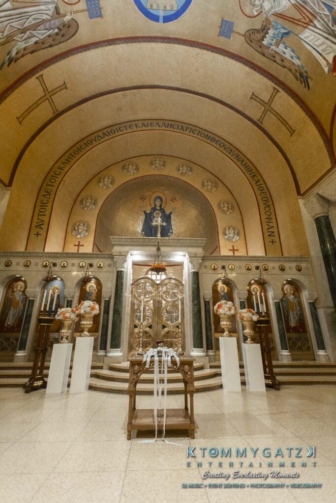 Ornate church interior with religious icons, Greek inscriptions, and decorated chairs. Center features a painting of a holy figure.