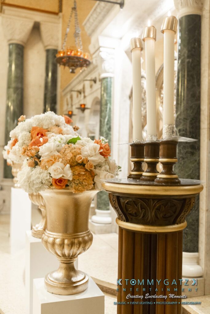 Floral arrangement with white and peach flowers in a gold vase beside a decorative stand holding candles, set in an ornate indoor setting.