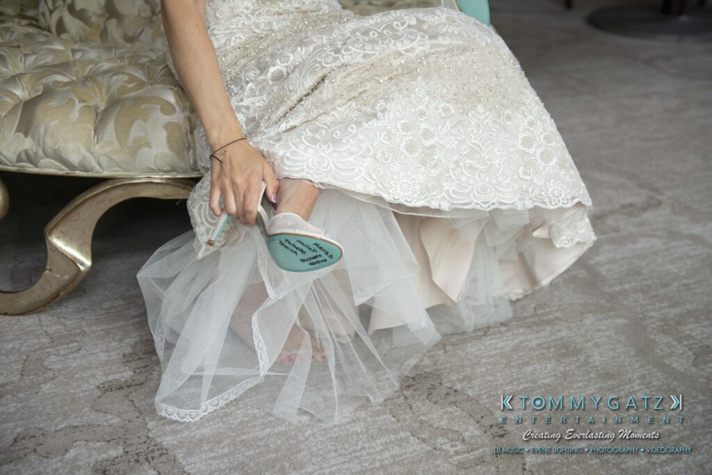 Bride sitting on a sofa, adjusting her shoe. The shoe soles display the message "I Do". Her lace wedding dress drapes gracefully around her.