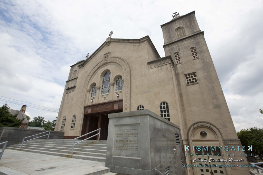 A beige church with twin towers and arched windows, featuring multiple crosses on its rooftop, situated against a cloudy sky. Steps lead up to the entrance.