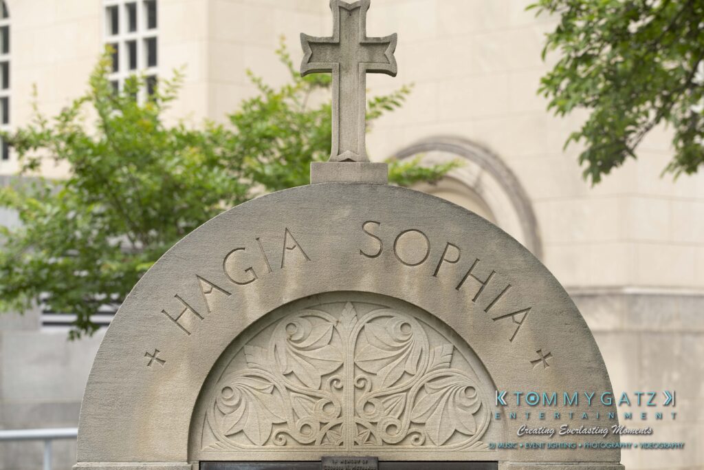 Stone archway with the words "Hagia Sophia" carved on it, featuring ornate floral designs and topped with a cross. Trees and a building are visible in the background.