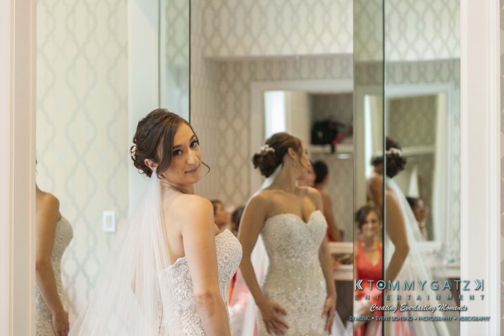Bride in a white dress stands in front of mirrors, adjusting her veil, with a reflection showing bridesmaids in the background.