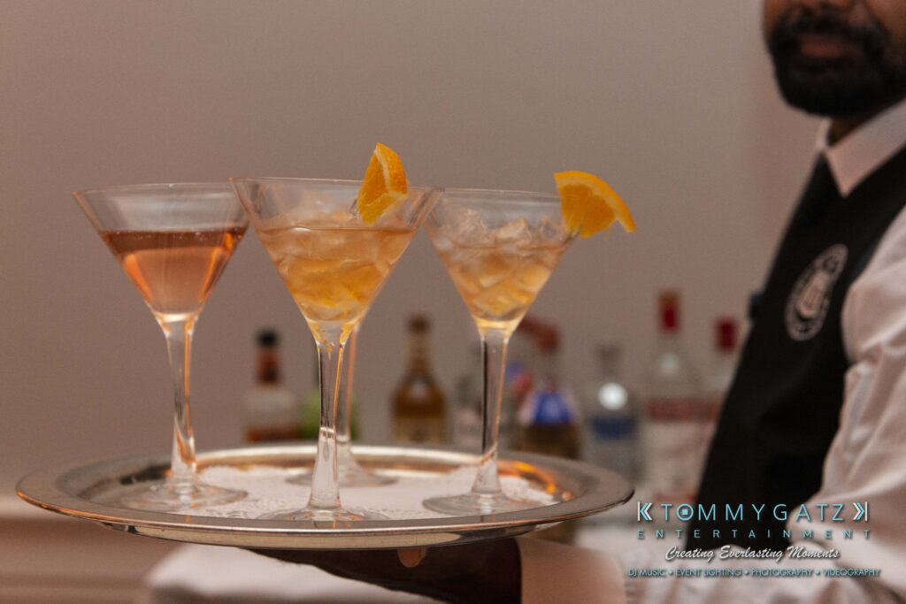 A waiter holds a tray with three martini glasses, each garnished with an orange slice, in front of a blurred background of liquor bottles.