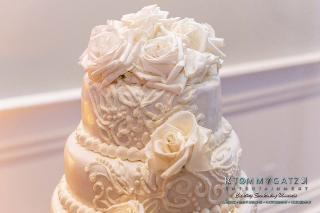 Three-tiered white wedding cake adorned with intricate frosting designs and topped with white roses.