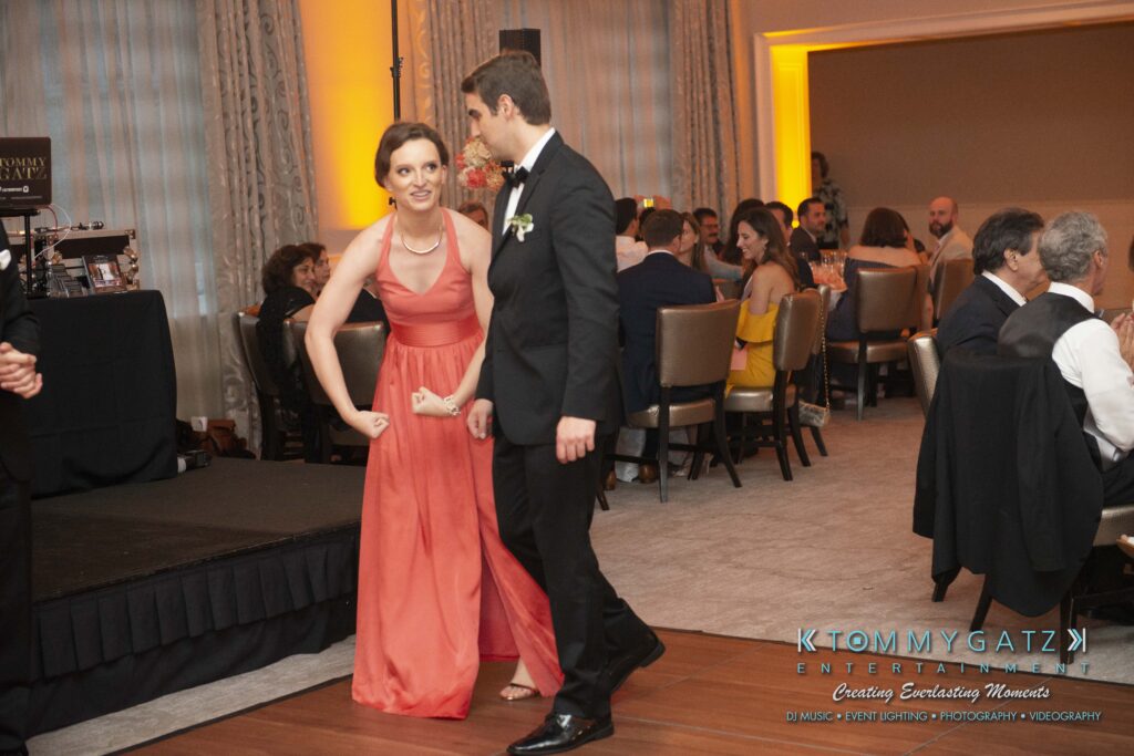 A woman in a coral dress strikes a playful pose on a dance floor next to a man in a suit, surrounded by seated guests at a formal event.